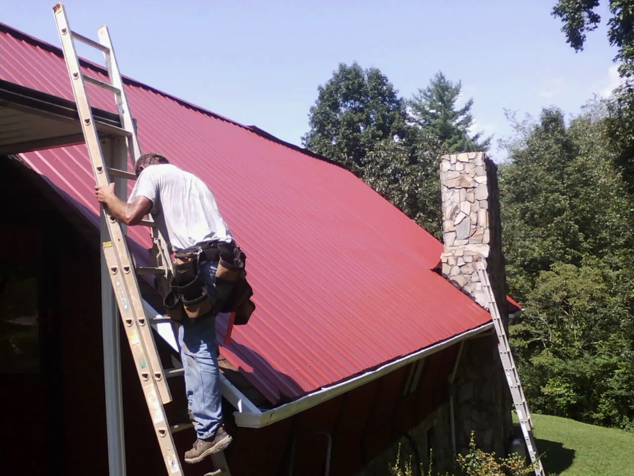 Roofer on a ladder beside a red metal roof and stone chimney — Soffit & Fascia Repair by Roofing Brothers in Peculiar